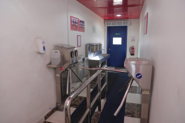 A hallway with a blue door, hand sanitizers, a sink, and a turnstile  The walls are white with red accents 