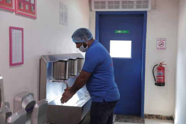 A man in a blue shirt and hairnet washes his hands at a stainless steel sink in a well lit hallway 