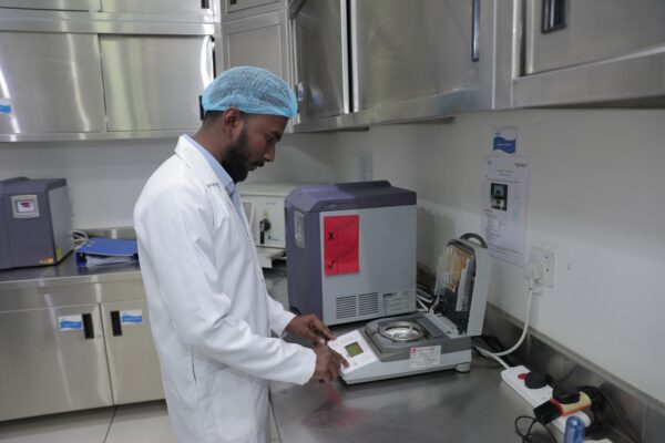 A person in a lab coat and hairnet operates a scientific instrument in a laboratory setting 