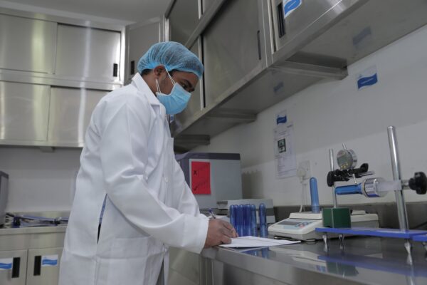 A person in a lab coat and mask is writing notes in a laboratory with equipment and blue test tubes in the background 