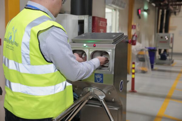 A person in a reflective safety vest is using a hand scanner at a turnstile in a facility 