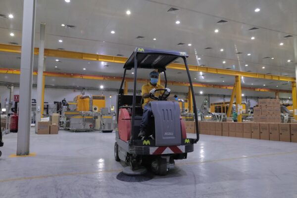 A person operates a floor sweeper in a large industrial space filled with machinery and stacked boxes 
