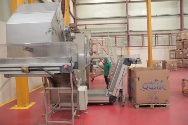 A worker in a green shirt operates machinery in a warehouse, with large boxes labeled "Oasis" in the background 