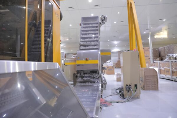 A conveyor system in a warehouse, featuring a metal incline and machinery, with boxes stacked in the background 