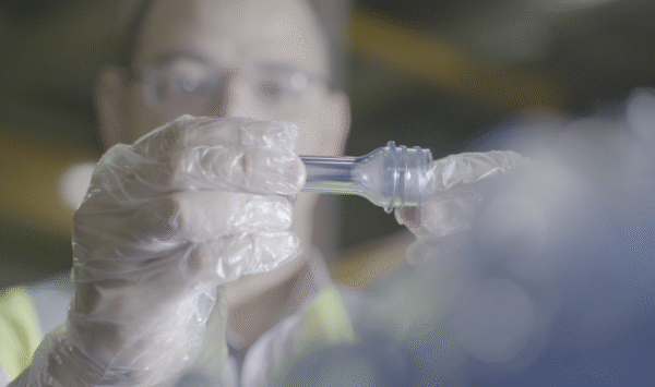 A person in gloves examines a clear plastic bottle component, focused on the item in a workshop setting 