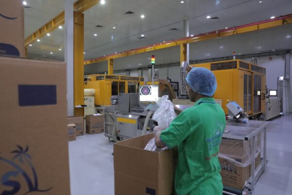 A worker in a green shirt is placing items into a large cardboard box in a factory setting with machinery in the background 